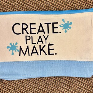May include: A rectangular canvas zipper pouch featuring a cream-colored upper section and a light blue lower section. The text "CREATE. PLAY. MAKE." is printed in black, accompanied by two light blue paint splatter graphics on the cream background.