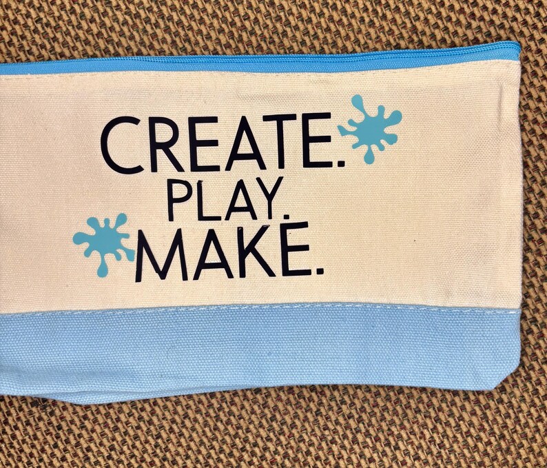 May include: A rectangular canvas zipper pouch featuring a cream-colored upper section and a light blue lower section. The text "CREATE. PLAY. MAKE." is printed in black, accompanied by two light blue paint splatter graphics on the cream background.