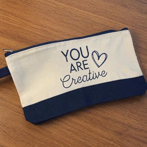 May include: A rectangular canvas zipper pouch featuring a two-tone design with a cream-colored upper section and a dark navy blue base. The text YOU ARE Creative is printed in dark navy blue, accompanied by a hand-drawn heart outline symbol.