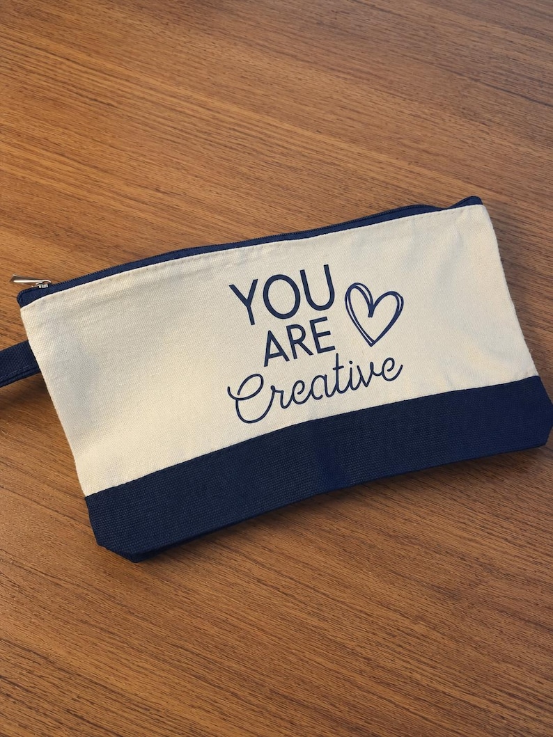May include: A rectangular canvas zipper pouch featuring a two-tone design with a cream-colored upper section and a dark navy blue base. The text YOU ARE Creative is printed in dark navy blue, accompanied by a hand-drawn heart outline symbol.