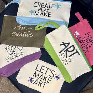 May include: A collection of six rectangular canvas zipper pouches in various colors including cream, olive green, purple, light blue, lime green, and pink. Each pouch features printed text such as CREATE. PLAY. MAKE., be creative, YOU ARE CREATIVE, ART IS FUN, HAPPY LITTLE ARTIST., and LET'S MAKE ART.