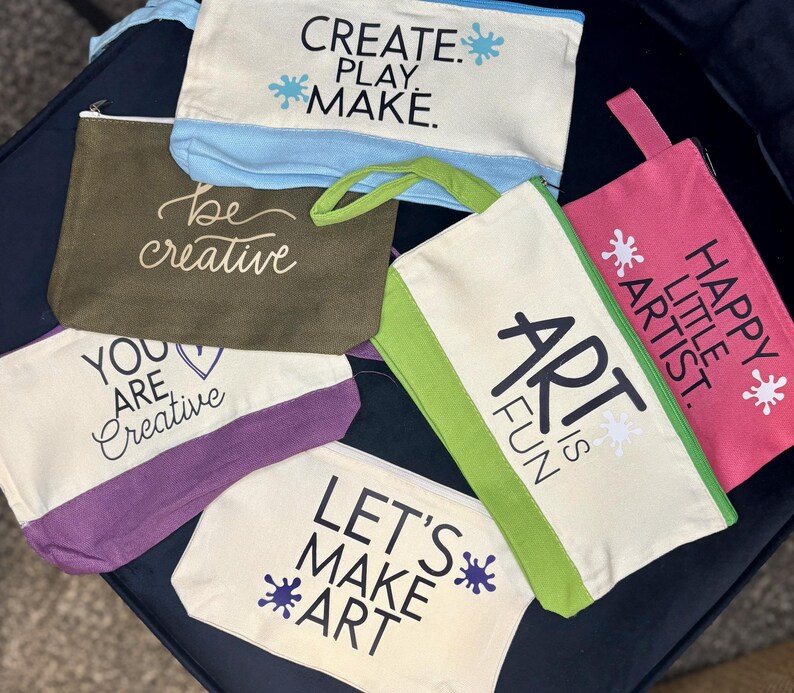 May include: A collection of six rectangular canvas zipper pouches in various colors including cream, olive green, purple, light blue, lime green, and pink. Each pouch features printed text such as CREATE. PLAY. MAKE., be creative, YOU ARE CREATIVE, ART IS FUN, HAPPY LITTLE ARTIST., and LET'S MAKE ART.