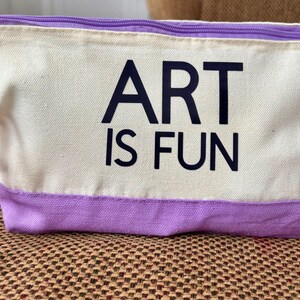 May include: A rectangular canvas pouch featuring a cream-colored upper section and a lavender-colored lower section. The text ART IS FUN is printed in black on the front. A lavender wrist strap is attached to the side with a purple metal carabiner.