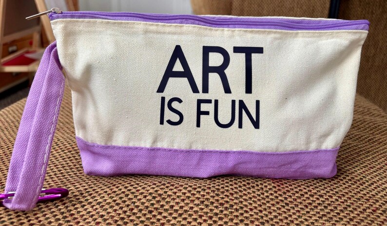May include: A rectangular canvas pouch featuring a cream-colored upper section and a lavender-colored lower section. The text ART IS FUN is printed in black on the front. A lavender wrist strap is attached to the side with a purple metal carabiner.