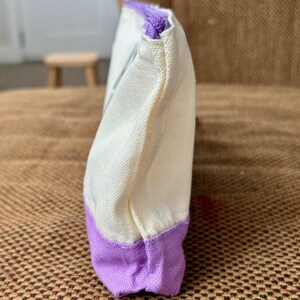 May include: A rectangular canvas pouch featuring a cream-colored upper section and a solid purple base. The pouch is constructed with a visible woven texture and includes a matching purple zipper closure along the top edge, viewed from the side.