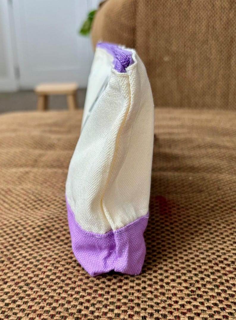 May include: A rectangular canvas pouch featuring a cream-colored upper section and a solid purple base. The pouch is constructed with a visible woven texture and includes a matching purple zipper closure along the top edge, viewed from the side.