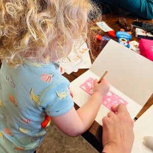 May include: A light blue t-shirt with a repeating pattern of yellow and pink sea creatures. The shirt is worn while using a pink plastic stencil with various shapes to draw on a white piece of paper with a wooden pencil on a wooden table.