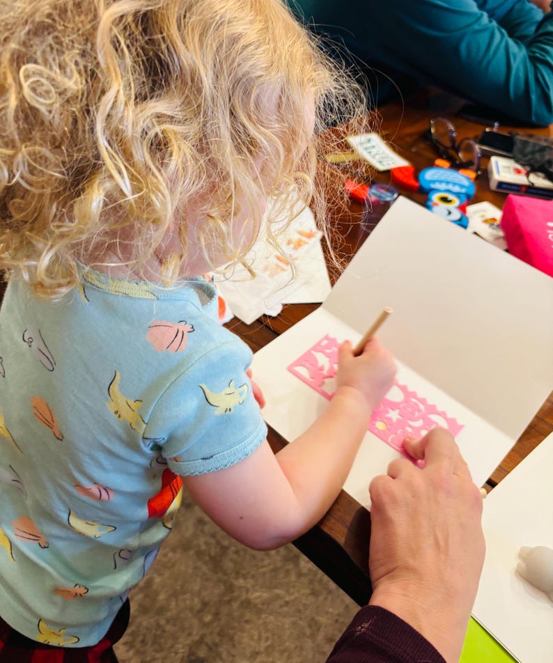 May include: A light blue t-shirt with a repeating pattern of yellow and pink sea creatures. The shirt is worn while using a pink plastic stencil with various shapes to draw on a white piece of paper with a wooden pencil on a wooden table.