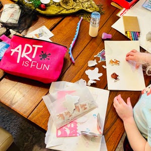 May include: A bright pink zippered pouch with the white text ART IS FUN and a black ink splatter graphic. The pouch is surrounded by various art supplies including a blue silicone fidget strip, a cylindrical container of colored pencils, and stickers.