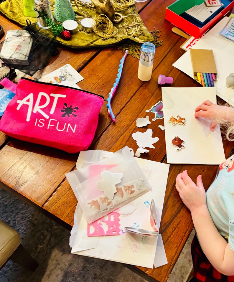 May include: A bright pink zippered pouch with the white text ART IS FUN and a black ink splatter graphic. The pouch is surrounded by various art supplies including a blue silicone fidget strip, a cylindrical container of colored pencils, and stickers.