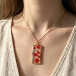 Puede incluir: Collar dorado con colgante rectangular. El colgante contiene flores rojas y pequeñas escamas doradas encapsuladas en resina transparente. El collar se lleva sobre una blusa color crema. El colgante mide 2,5 cm x 1,5 cm.