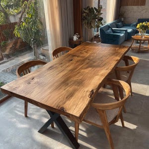 May include: A large, rectangular wooden dining table with a natural, live edge. The table has a dark brown finish and is supported by a black metal base. Four wooden chairs with curved backs surround the table. The room has large windows and a modern design.