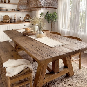 Puede incluir: Una mesa de comedor rústica de madera con un banco y sillas a juego. La mesa tiene un acabado envejecido y está decorada con un camino de mesa blanco, jarrones y una bandeja tejida. La habitación tiene una paleta de colores neutros.
