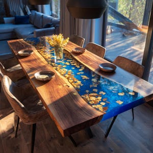 May include: A handcrafted wooden dining table with a blue epoxy river and embedded golden leaves. The table is surrounded by brown leather chairs and has a vase of yellow flowers. The table is approximately 96 inches long.