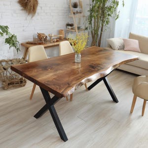Könnte beinhalten: Ein großer, rechteckiger Esstisch aus Holz mit natürlicher Kante. Die Tischplatte hat eine satte braune Farbe und wird von schwarzen Metallkreuzbeinen getragen. Der Tisch ist von Stühlen und einem Sofa umgeben, was auf eine Wohnzimmereinrichtung hindeutet.