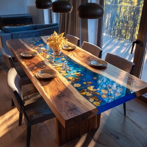 May include: A handcrafted wooden dining table with a blue epoxy resin river design. The table features a live edge and is adorned with embedded golden leaves. Includes six matching chairs and decorative plates. The table measures approximately 96 inches long.