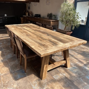 Puede incluir: Una gran mesa de comedor rectangular de madera con acabado natural. La mesa tiene una base de caballete resistente y está rodeada de varias sillas de madera con asientos de cuero. La mesa mide aproximadamente 3 metros de largo.