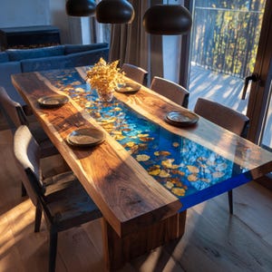 May include: A handcrafted wooden dining table with a blue resin river design. The table features embedded golden leaves and is surrounded by chairs and place settings. The table is approximately 100 inches long.