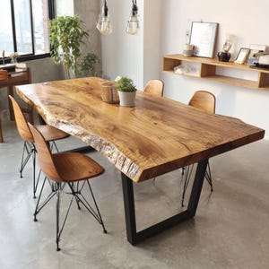 Könnte beinhalten: Ein großer, rechteckiger Esstisch aus Holz mit einer Baumkante und einem schwarzen Metallgestell. Vier Holzstühle mit schwarzen Drahtbeinen umgeben den Tisch. Eine kleine Topfpflanze und dekorative Gegenstände stehen auf dem Tisch.