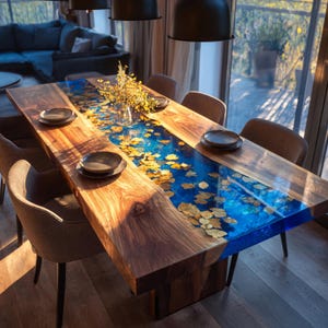 May include: A large wooden dining table with a blue resin river design, inlaid with gold-colored leaf accents. The table is surrounded by six brown upholstered chairs and set with dark plates. A vase of yellow flowers sits in the center.