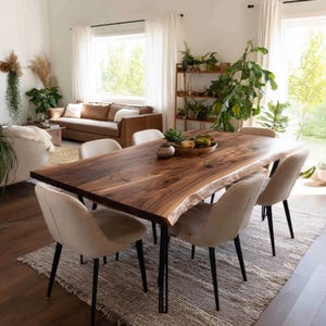 Op de afbeelding: Een houten eettafel met een natuurlijke rand, gecombineerd met zes beige gestoffeerde stoelen. De tafel, met zijn natuurlijke houtnerf, staat op zwarte metalen poten. De stoelen zijn rond de rechthoekige tafel geplaatst, die op een patroonkleed staat. De kamer heeft planten en een bruine leren bank.