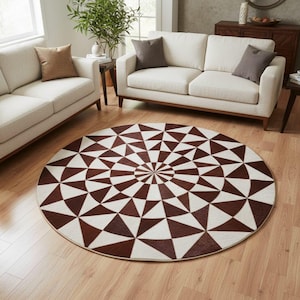 Może przedstawiać: Okrągły dywan z geometrycznym wzorem w odcieniach brązu i kremu. Dywan ma radialny wzór trójkątów, tworząc uderzający efekt wizualny. Dywan jest umieszczony na drewnianej podłodze, uzupełniając neutralne tony pomieszczenia. Średnica około 200 cm.