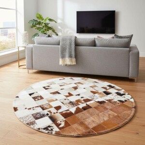 May include: A circular rug with a patchwork design. The rug features squares of white, brown, and black cowhide. The rug is on a wooden floor in a living room setting. A grey sofa and a television are in the background.