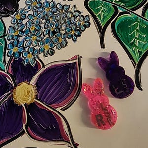 May include: A vibrant image featuring a floral design with purple, pink, and green elements. Two glittery bunny-shaped resin charms, one pink and one purple, are also present. The background is white.