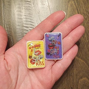 Pode incluir: Dois pingentes retangulares de resina com temas românticos são segurados em uma mão. Um pingente é amarelo com batom e as palavras "Love", "Kiss" e "XOXO". O outro é roxo com "XOXO", um batom e a palavra "Love".