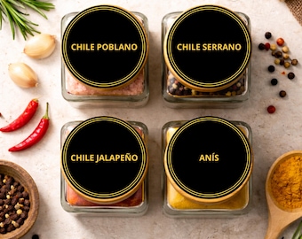 May include: Four square glass spice jars with black circular labels and gold trim. The labels read "CHILE POBLANO", "CHILE SERRANO", "CHILE JALAPEÑO", and "ANÍS". Red chili peppers, garlic cloves, and spices are also visible.