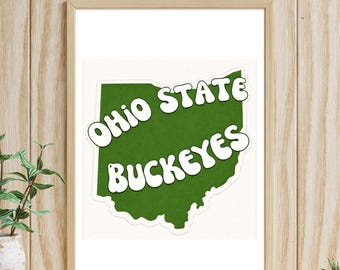 May include: A framed print featuring a green outline of Ohio with the words "Ohio State Buckeyes" in white, retro-style lettering. The print is set against a white background and framed in a light wood-tone frame.