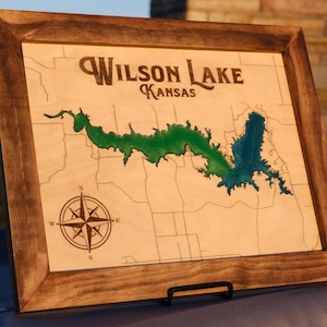 Könnte beinhalten: Holzgerahmte Karte des Wilson Lake, Kansas, mit der Umrisslinie des Sees in Grün und Blau. Die Karte enthält eine Windrose und den Text "Wilson Lake Kansas" in einer dekorativen Schriftart. Der Rahmen ist dunkelbraun.