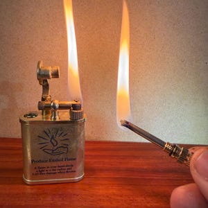May include: A brass-colored lighter with a flame, next to a lit match. The lighter has the text "Produce Exalted Flame" and a graphic. The flame is orange and white. The lighter is on a wooden surface.