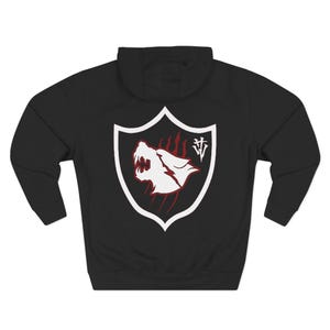 May include: Black hooded sweatshirt featuring a white shield design with a snarling wolf head and red claw marks. The shield also includes a white monogram. The sweatshirt has a drawstring hood and long sleeves.