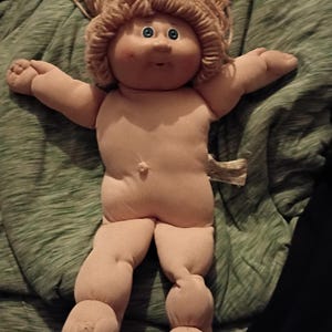 May include: A vintage Cabbage Patch Kid doll with a soft, fabric body and a vinyl head. The doll has light brown yarn hair, blue eyes, and rosy cheeks. It is wearing no clothes and has its arms outstretched.