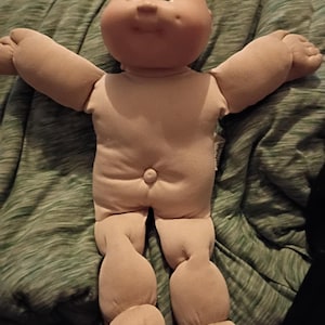 May include: A tan-coloured doll with a round head, blue eyes, and a small nose and mouth. The doll's arms are outstretched, and its legs are bent. The doll is made of soft fabric and has a simple, classic design. The doll is lying on a green blanket.