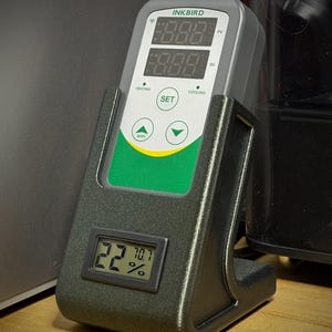 May include: A gray and black Inkbird digital temperature and humidity controller. The device displays temperature and humidity readings on its digital display. The device is in a black stand and has buttons for setting and controlling the temperature.
