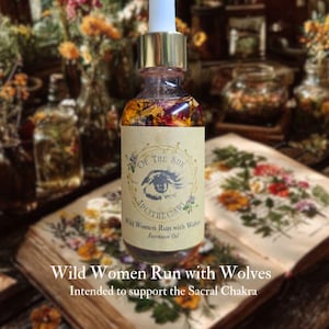 May include: A glass bottle of intention oil with a dropper, filled with dried flowers and herbs. The label reads "Wild Women Run with Wolves" and "Intended to support the Sacral Chakra." The bottle sits on an open book with floral illustrations.