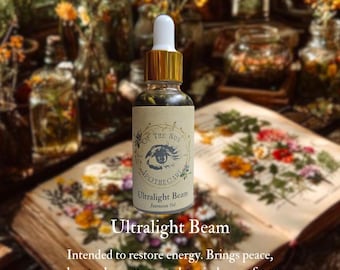 Puede incluir: Una botella de vidrio transparente con tapa dorada y cuentagotas blanco, etiquetada como "Ultralight Beam Intention Oil" de "Of The Sun Apothecary". La botella está sobre un libro abierto con ilustraciones florales.