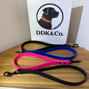 May include: Three dog leashes in black, blue, and pink are displayed on a wooden surface. Each leash has a loop handle and a black metal clasp. A white sign with a black dog silhouette and the text "DDK&Co." is in the background.