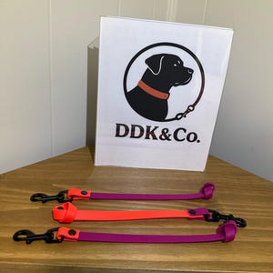 May include: Three dog leashes in vibrant colors: purple, orange, and purple. Each leash has a black clasp and a matching accent piece. A white sign with a black dog logo and the text "DDK&Co." is in the background.
