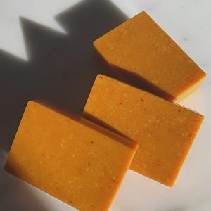 May include: Three rectangular bars of orange soap are arranged on a white surface. The soap has a textured appearance with small, reddish specks. A shadow from an unseen object casts a dark shape across the top left of the image.