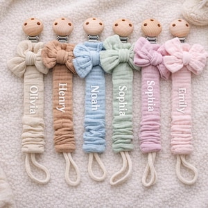 May include: Six pastel-coloured baby pacifier clips, each with a bow and a name printed on the fabric. The clips have wooden details and are arranged on a soft, textured surface.