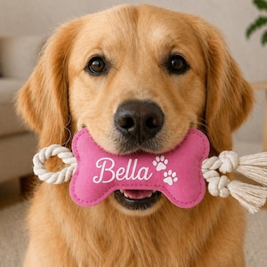 May include: A golden retriever with a pink bone-shaped dog toy in its mouth. The toy has the name "Bella" in white script and two paw prints. The toy is attached to a white rope.