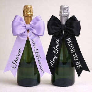 Peut inclure: Deux bouteilles de champagne décorées de nœuds et de rubans. L'une avec un nœud lilas et un ruban « Sharon Happy 50th Birthday », l'autre avec un nœud noir et « Amy Smith Bride to Be ».