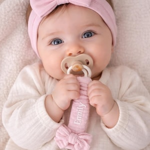 May include: A light pink ribbed headband with a bow, a beige pacifier, and a matching light pink ribbed pacifier clip featuring the name Emily embroidered in white thread, a small fabric bow, and a round wooden clip with three ventilation holes.