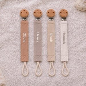 Könnte beinhalten: Vier Schnullerketten aus Stoff in den Farben Terrakotta, Taupe, Beige und Creme. Jede Kette hat oben einen runden Holzclip und unten eine weiße Kordelschlaufe. Die Namen Olivia, Henry, Noah und Hannah sind in weißer Schrift aufgestickt.