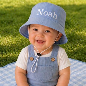 May include: A baby wearing a pale blue bucket hat with the name "Noah" printed on it. The baby is also wearing a denim blue dungarees and a white t-shirt. The baby is smiling and sitting on a blue and white checked blanket.