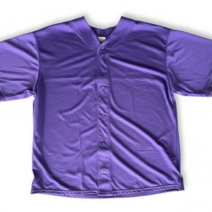 Puede incluir: Una camiseta de béisbol morada lisa con mangas cortas y cierre de botones. La camiseta tiene cuello en V y está hecha de una tela suave y ligera. La camisa está tendida sobre una superficie blanca.