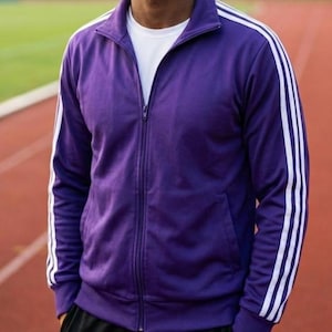 May include: A purple track jacket with white stripes down the sleeves. The jacket features a full zip, a stand-up collar, and a white t-shirt underneath. The jacket is made of a smooth, athletic material.